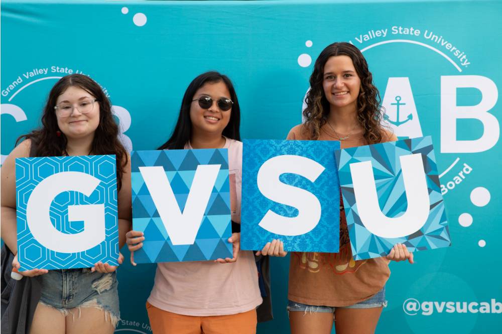 three students posing in front of CAB backdrop at Laker Kickoff photo booth and holding GVSU letters